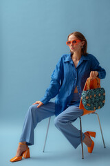Fashionable young woman wearing trendy orange sunglasses, blue linen shirt, trousers, block heel shoes, holding straw wicker bag, posing on blue background. Studio portrait. Copy, empty space for text - 650386160
