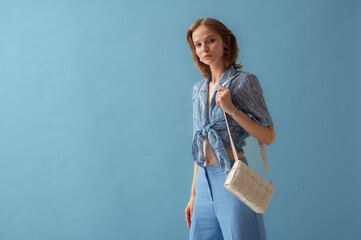 Fashionable young woman wearing  knotted chiffon blouse, trousers, carrying trendy white leather padded cassette shoulder bag, posing on blue background. Studio portrait. Copy, empty space for text - 650386169