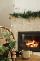Cozy winter. Stylish cup of warm tea, basket with fir branches, wooden trees and star, pine cones on table against burning fireplace. Modern rustic eco friendly decor in scandinavian room