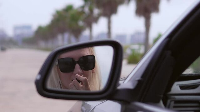 Beautiful woman with blonde hair driving car paints her lips with lipstick. Woman driving doing makeup. Reflection of woman in rearview mirror. Successful women in car