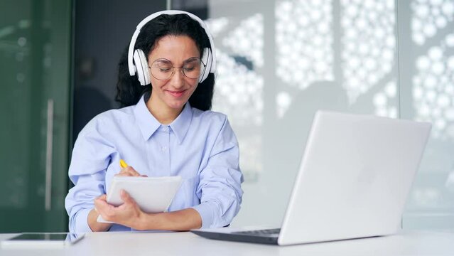 Female employee in headphones listens to online e-learning, video course sitting in office. Woman at a distance learning seminar writes in a notebook using a laptop computer communicates with a coach