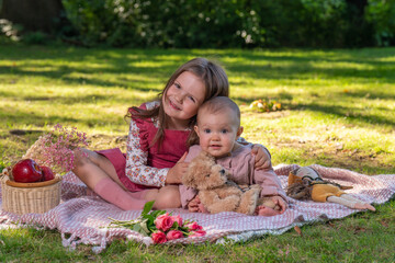 Portrait of cute girls 5 years old and 1 year old. Two sisters on a picnic walk. They look fun at the camera. There is a teddy bear nearby and red apples in a basket.