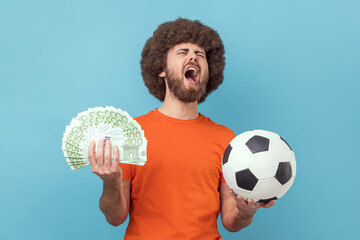 Excited man with Afro hairstyle wearing orange T-shirt holding soccer ball and hundred euro bills,...
