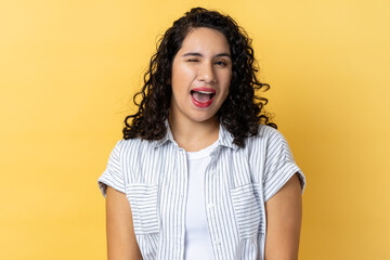 Portrait of cheerful attractive woman with dark wavy hair being in good mood, smiling broadly and winking at camera with toothy smile. Indoor studio shot isolated on yellow background. - obrazy, fototapety, plakaty