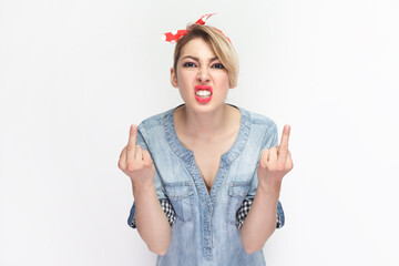 Portrait of angry aggressive rude blonde woman wearing blue denim shirt and red headband standing showing fuck sign, arguing with somebody. Indoor studio shot isolated on gray background. - obrazy, fototapety, plakaty
