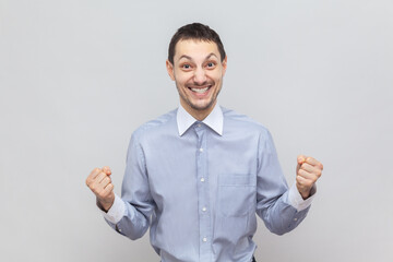 Portrait of extremely happy joyful cheerful man standing with clenched fists, rejoices his success, celebrating victory, wearing light blue shirt. Indoor studio shot isolated on gray background.