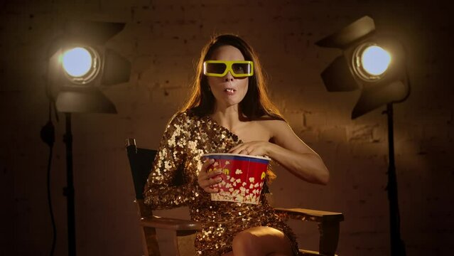Woman in golden dress and paper glasses sitting on a director's chair, watching scary movie, eating popcorn and flinches, drops popcorn.