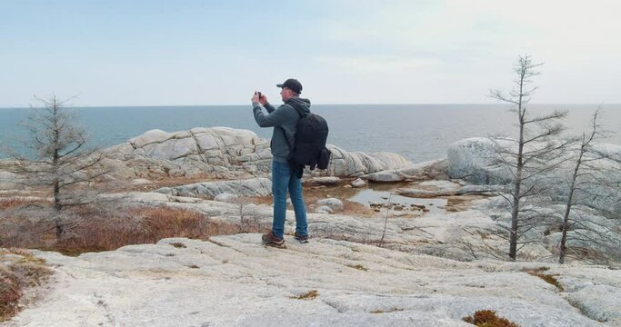 Men on a Rocky Shore with a Rucksack Uses Smartphone to Photograph the Landscape