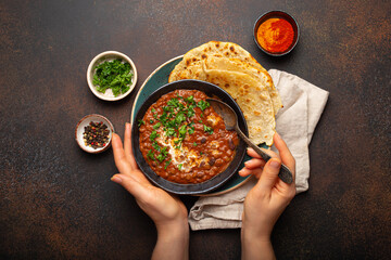 Female hands holding a bowl and eating traditional Indian Punjabi dish Dal makhani with lentils and beans served with naan flat bread, fresh cilantro on brown concrete rustic table top view.