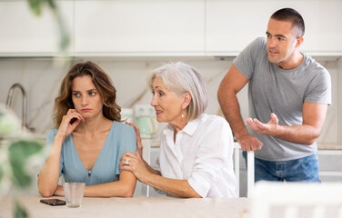 Fototapeta premium Young girl is sitting at table in kitchen quarreling with her mother-in-law and husband
