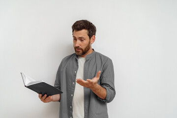Baffled man in casual clothes standing with open notepad and looking on it on white background