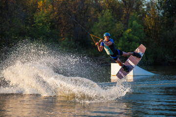 An athlete does a trick on the water. A rider jumps on a wakeboard against a background of a green forest. Sunset on the lake