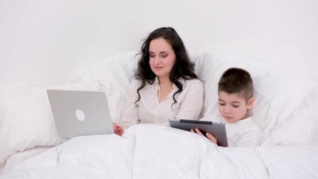 mom and son under blanket playing on tablet boy 4-6 years old woman with black hair beautiful lies under white sheet press buttons laugh smile games internet. online learning educational activities