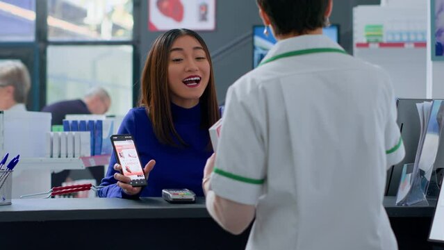 Asian customer at drugstore counter, showing druggist prescribed cardio medicine picture on smartphone, ready to purchase it. Wellness expert selling client pain relieving pills