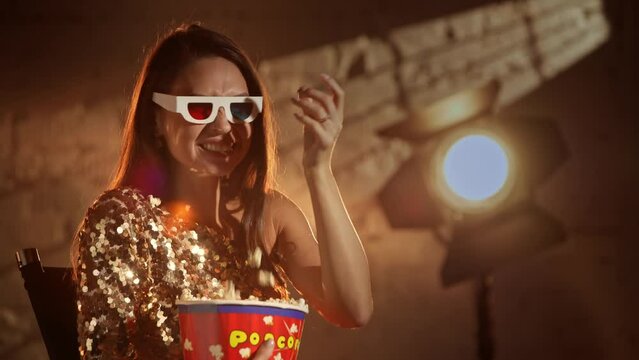 Closeup shot of attractive woman in golden dress wearing 3d paper glasses, watching comedy laughing and eating popcorn from bucket.