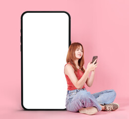 Young smiling happy Asian woman in red shirt sit near big mobile cell phone or smartphone with blank screen workspace area chatting isolated on pink background.