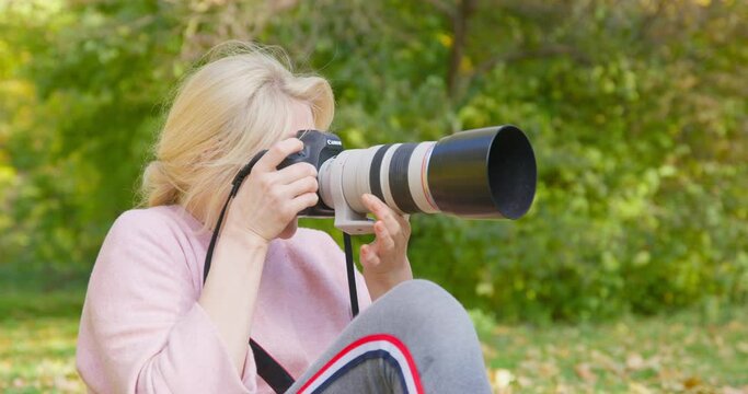 Young blonde woman is photographing something in park or forest using camera with large telescopic lens. She zooms in and out of focus to catch the best shot of nature