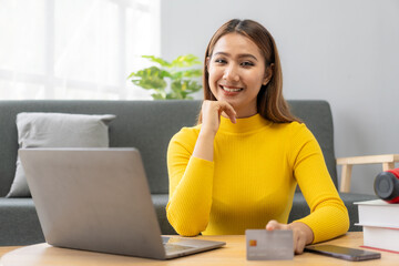 Asian girl pays online using laptop to shop at home Attractive Asian millennial woman holds her smartphone and credit card using mobile banking or online shopping app.