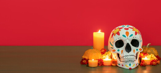 Painted human skull for Mexico's Day of the Dead (El Dia de Muertos), candles, flowers and pumpkins on red background with space for text