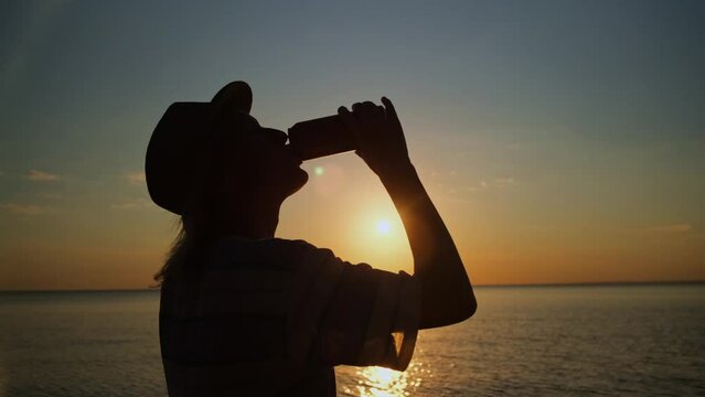 Traveler woman silhouette drinking beer from tin can lit by sunset beams overlooking sea. Alcohol relaxation in vacation.