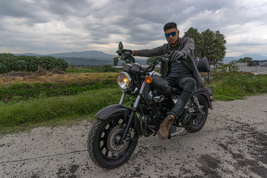 Hombre negro con lentes posando en su moto en carretera solitaria y bello paisaje verde de fondo. 