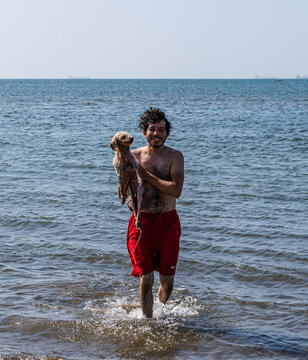 Hombre joven en traje de ba&ntilde;o, feliz cargando a su perro en la orilla de la playa, disfrutando de una hermosa tarde calurosa en compa&ntilde;&iacute;a de su mejor amigo.