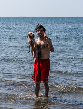 Chico adolescente en la playa cargando a su perro, ambos felices disfrutando un d&iacute;a en la playa con el inmenso mar azul al fondo.