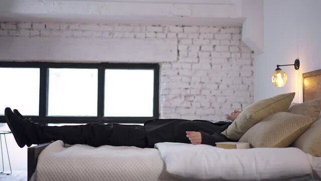 Side view happy tired man in suit jumping up falling back on bed in slow motion. Wide shot satisfied Caucasian freelancer businessman in bedroom at home indoors