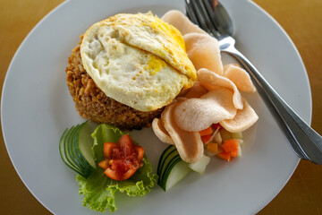 Javanese fried rice consisting of rice, fried eggs, pickled cucumber, vegetables on a white plate with cutlery which is a menu in a cafe and restaurant.