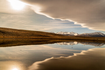A small pool of water on rural farming fields reflecting the snow covered southern alps and a n amazing cloudscape
