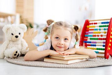 a child girl reads a book lying on the floor among toys in a bright bright nursery or kindergarten, the concept of children's learning centers or kindergartens and learning