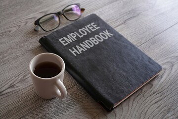 Closeup image of book with text EMPLOYEE HANDBOOK surrounded by cup of coffee and glasses on office...