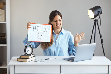 Young and positive teacher greets an online student on a laptop and holds a sign that says hello....