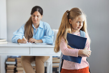  Little schoolgirl is worried about a grade on a test at school, a teacher checks the exam of schoolchildren in the background. Experiences and emotions in education concept