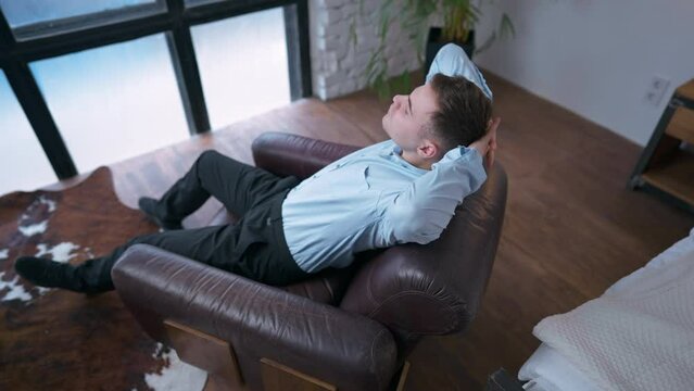 Wide shot relaxed tired businessman sitting in armchair placing hands behind head in slow motion. High angle view portrait of exhausted satisfied Caucasian young man at home indoors