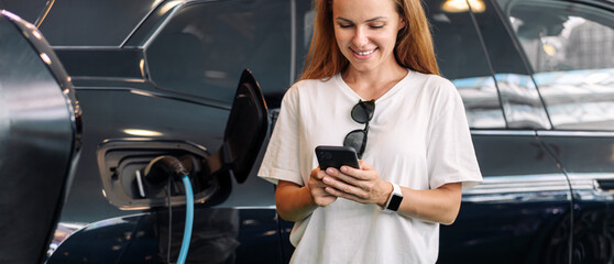 Young woman standing in front of electric electric vehicle smiling and using her mobile phone....