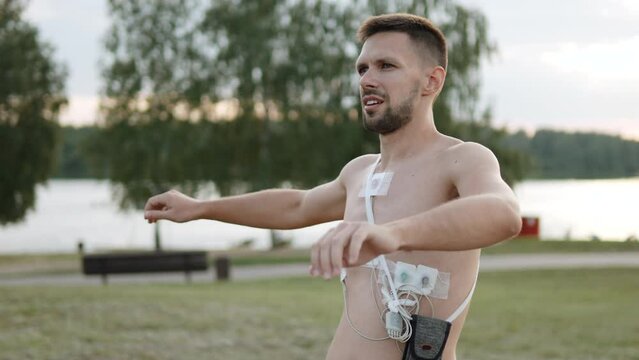 Slender guy wearing holter monitor to study work of heart does exercises in park, arc shot. Man makes jerks with his arms with sensors glued to record his heart rate on his bare chest.