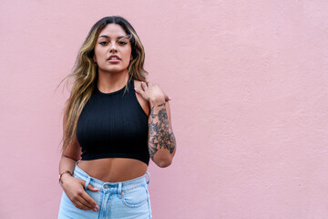 Casually dressed, this beautiful young Latin woman showcases her tattoos against a vibrant pink...