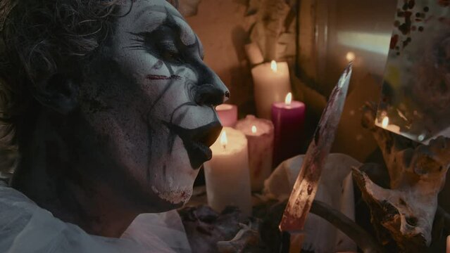 Close up of mad clown with scary makeup bringing knife to candle flame and dragging it across face, sitting in front of mirror
