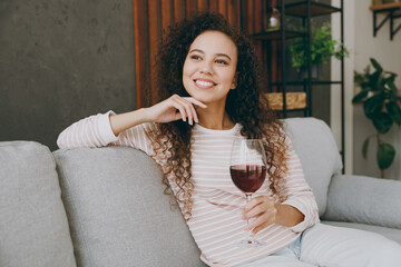 Side view young woman wear casual clothes sits on grey sofa couch look aside prop up chin drinking wine stay at home hotel flat rest relax spend free spare time in living room indoor. Lounge concept.