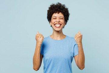 Young fun woman of African American ethnicity wear t-shirt casual clothes doing winner gesture celebrate clenching fists say yes isolated on plain pastel light blue cyan background. Lifestyle concept.