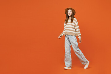 Full body side profile view young woman she wearing striped sweater hat casual clothes walking going look camera isolated on plain orange red color wall background studio portrait. Lifestyle concept.