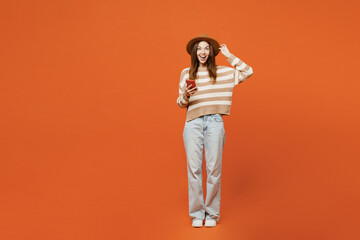 Full body surprised excited young woman wear striped sweater hat casual clothes hold head use mobile cell phone isolated on plain orange red color wall background studio portrait. Lifestyle concept. - obrazy, fototapety, plakaty