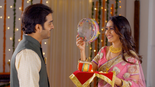 Young Indian couple happily performing Karwa Chauth traditions in traditional wear. Beautiful wife looking at the moon through a sieve and then to her good looking husband  following Karwa Chauth r...