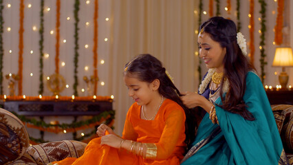 South Indian mother wearing traditional dress is helping her daughter with hairstyle - Festival at Home. Beautiful mother making braid hairstyle for her cute daughter while she is busy playing with...