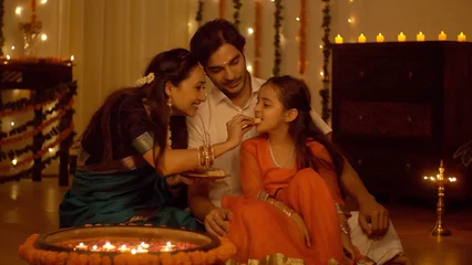 Fotobehang South Indian family dressed in traditional wear is celebrating Diwali at home - Happy Family. Family spending time together  eating sweets and celebrating Diwali - Festive colorful background with ... © Knot9 Images
