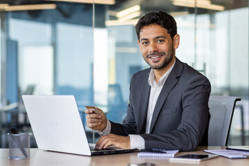 Fototapeta na wymiar Portrait of young hispanic businessman inside office at workplace, man holding bank credit card in hands, using laptop for online shopping and money transfer, employee smiling and looking at camera.