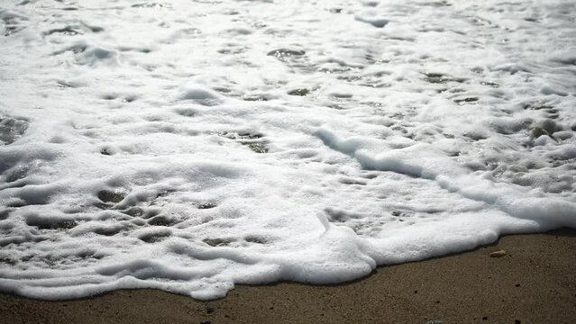 Closeup footage of foamy sea waves wahing off footprints on the golden sand at beach. Close-up 4k video in sunny weather.
