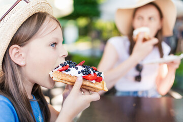 Cute little girl eating a piece of waffles with whipped cream, strawberries and blueberries with her sister - 650596724