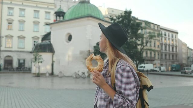 Happy young blonde female tourist with stylish clothes and a boho hat eating bagel obwarzanek traditional polish cuisine snack waling on Market square in Krakow. Traveling Europe. St. Marys Basilica